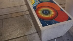 Una larga mesa con mosaico colorido y cubierta de vidrio, rodeada de sillas blancas en un espacio público.