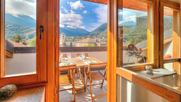 A wooden balcony with a table set for dining, large windows with mountain views, and a stone on the floor. - Sunny Penthouse With Mountain View (Oulx)