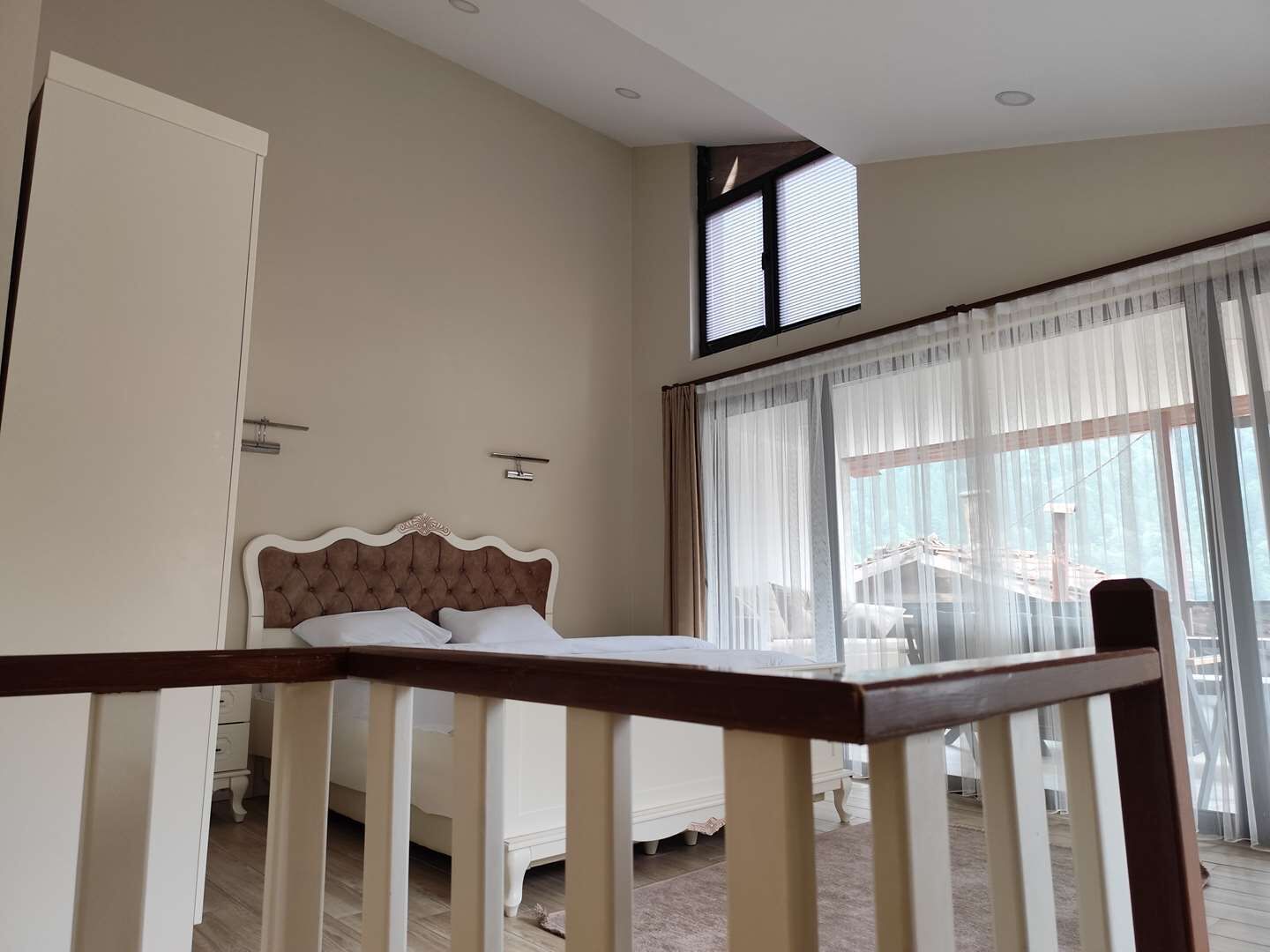 Une chambre à coucher avec un lit, une fenêtre avec des stores, une porte et une rambarde d’escalier.