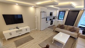 A modern living room with a flat-screen TV, a white sofa, a coffee table, and a kitchen area with white cabinets and a window. - Garden House Uzungöl (Çaykara)