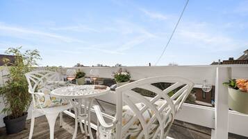 Terraço no telhado com mesa redonda e cadeiras brancas, rodeado por plantas em vasos e céu azul límpido.