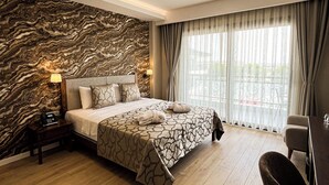 A hotel room with a marble-patterned wall, a large bed with patterned bedding, a bedside table with a phone, and a view of the outdoors through sheer curtains. - Dedeman Gure Nurhayat Thermal Resort&Spa (Edremit)