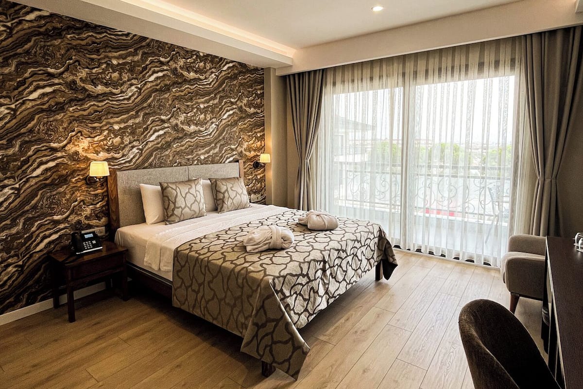 Une chambre d’hôtel avec un mur décoré d’un motif imitant le marbre, un grand lit avec une literie à motifs, une table de chevet avec un téléphone et une vue sur l’extérieur à travers des rideaux transparents.