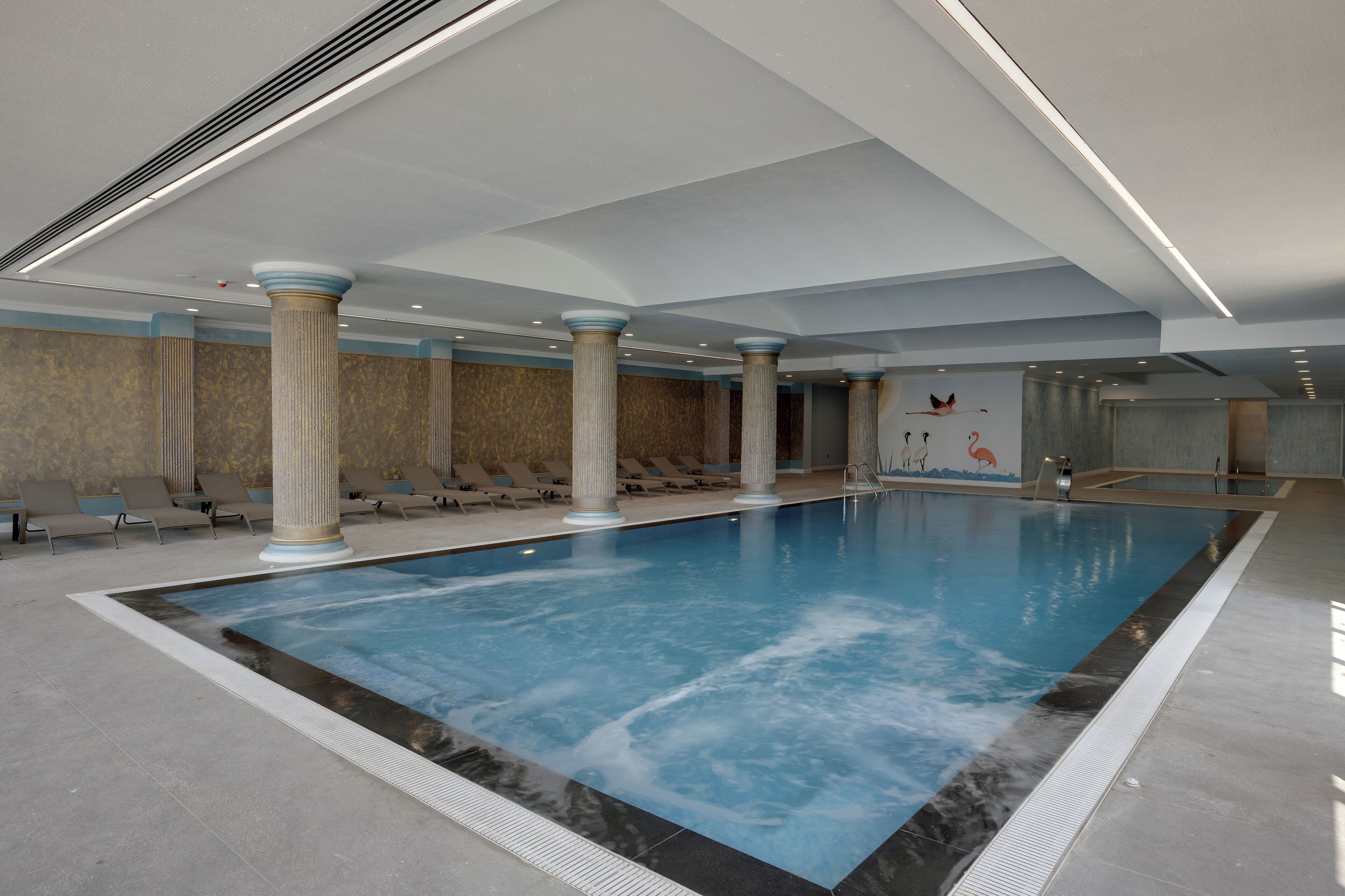 Ein großes Hallenbad mit Säulen, Liegestühlen und einem Wandgemälde.