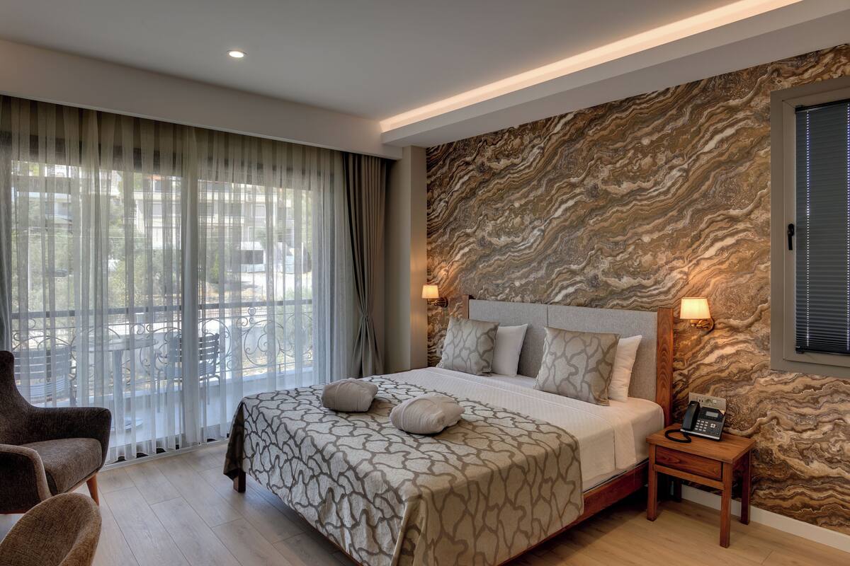 Une chambre d’hôtel moderne dotée d’un grand lit, d’un coin salon avec des fauteuils, d’un mur décoratif en pierre et d’une vue dégagée à travers des rideaux légers.