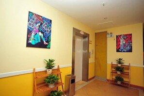 Corredor de hotel com um banco de madeira, plantas em vasos e duas pinturas nas paredes.