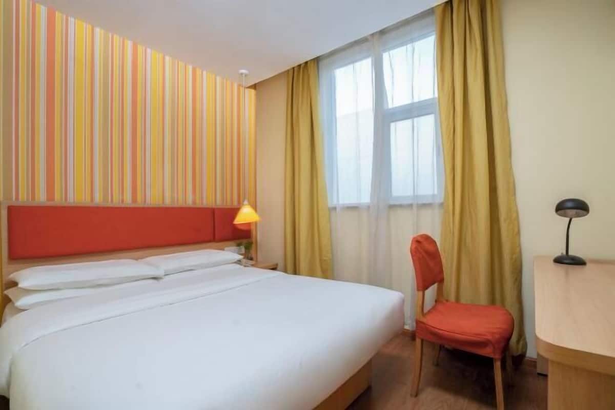A hotel room with a bed, a chair, a desk, a lamp, a window with curtains, and striped wall decor.