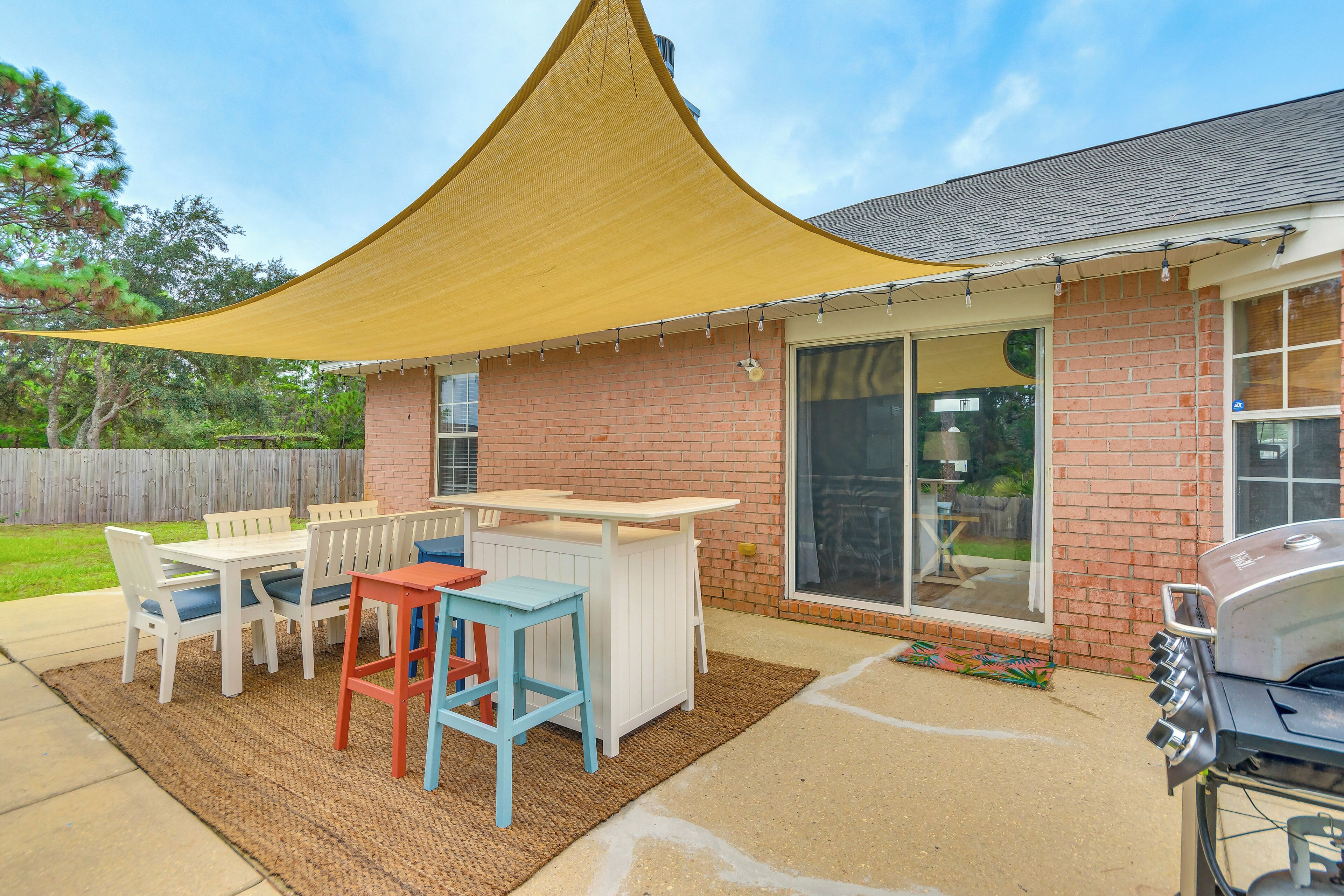 A backyard with a barbecue grill, a white table and chairs, and a yellow shade sail.