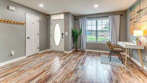 A spacious room with hardwood flooring, a white desk, a chair, a potted plant, and a large window with curtains. - Pensacola Home w/ Fire Pit, 7 Mi to Beaches! (Pensacola)
