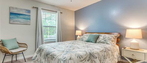 A bedroom with a bed, a ceiling fan, a nightstand with a lamp, a chair, a window with blinds, and a painting on the wall.