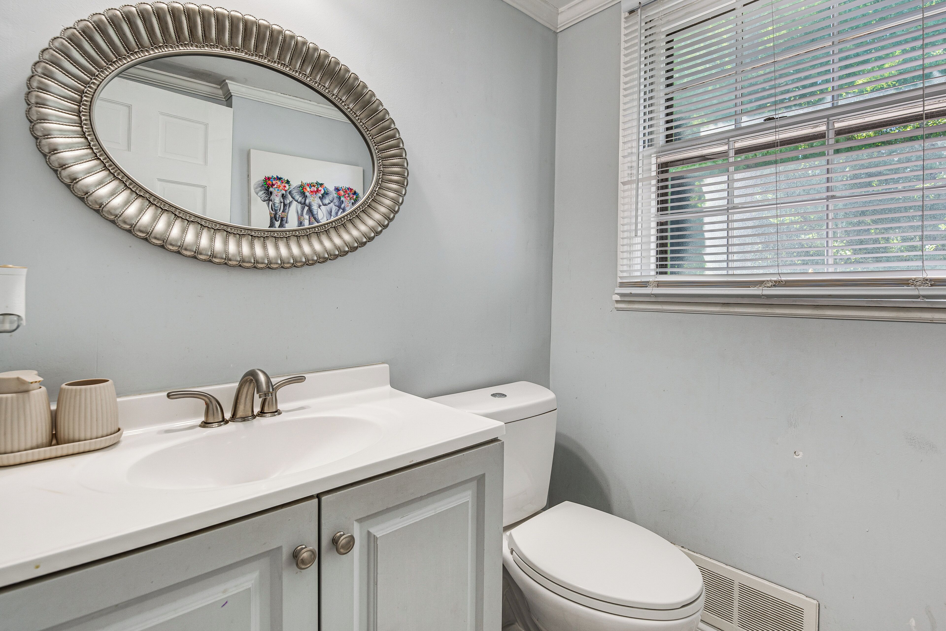 A bathroom with a white sink, a toilet, a mirror with a decorative frame, and a window with blinds.