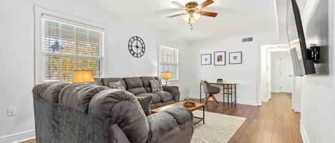 A living room with a grey sofa, a ceiling fan, and a flat-screen TV mounted on the wall.