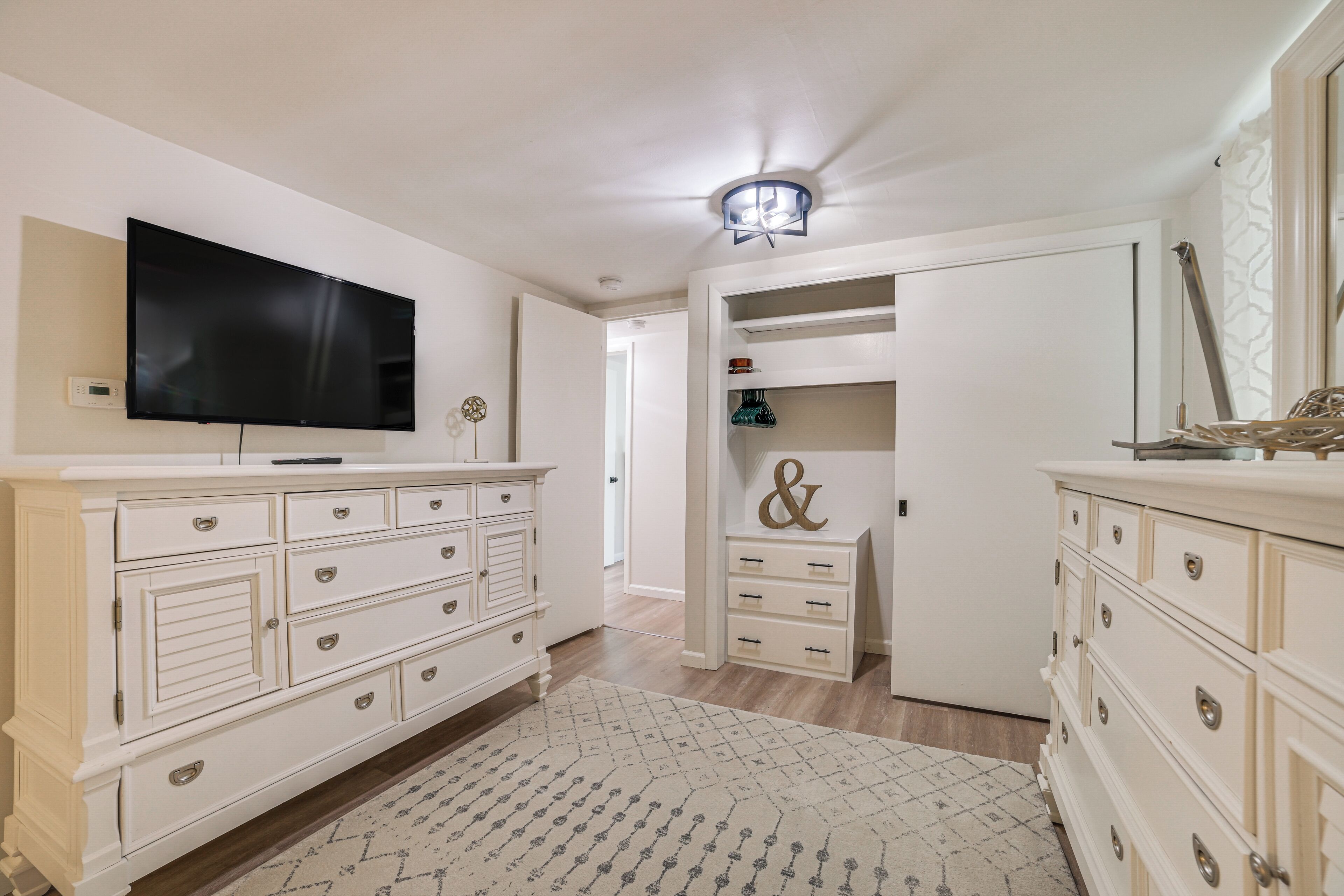 Une chambre avec une télévision, des meubles blancs, un luminaire au plafond et un placard.