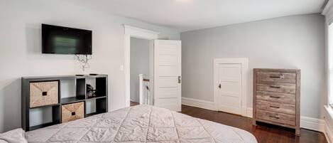 A bedroom with a bed, a TV mounted on the wall, a dresser, and a door leading to another room.