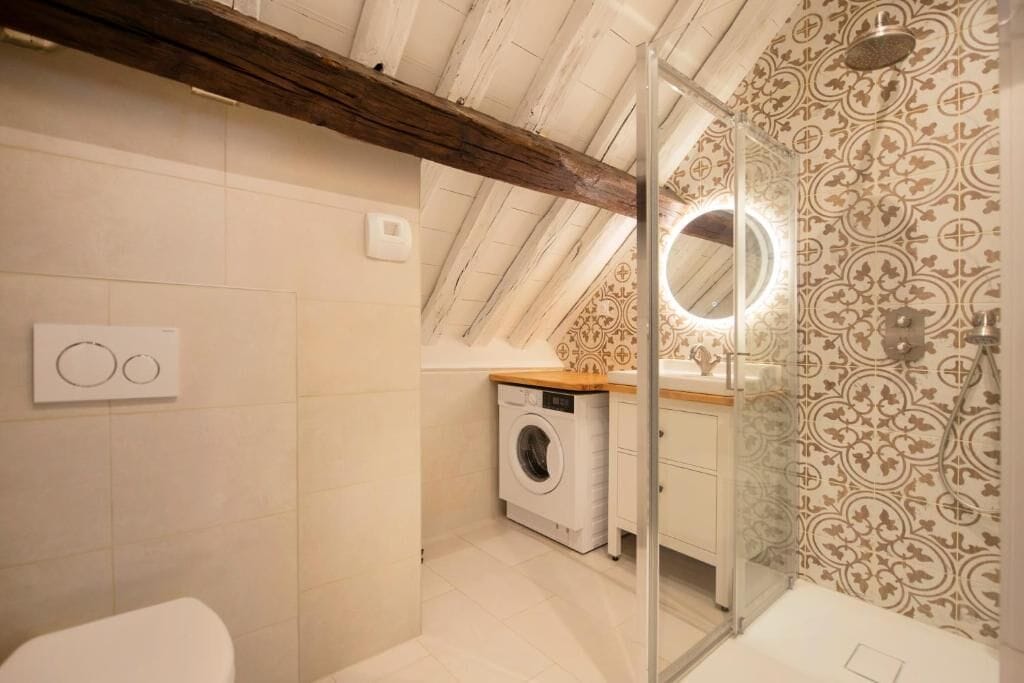 A compact bathroom with a washing machine, a glass shower enclosure, and a wooden ceiling beam.