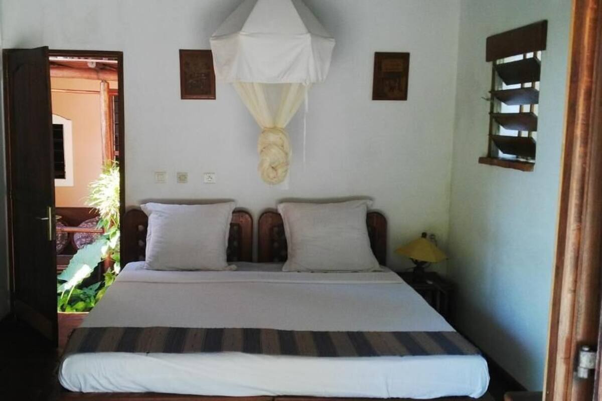 Un dormitorio con cama, mosquitera, mesita de noche y ventana con estante.