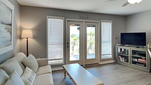 A living room with a sofa, a coffee table, a TV on a cabinet, and a ceiling fan. - 1BD/1BA Condo w. Hall Bunk Bed, Steps from Beach, Pool + Hot Tub (Perdido Key)