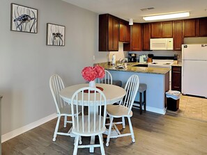 A modern kitchen with wooden cabinets, a white refrigerator, and a dining area with white chairs and a vase of flowers.