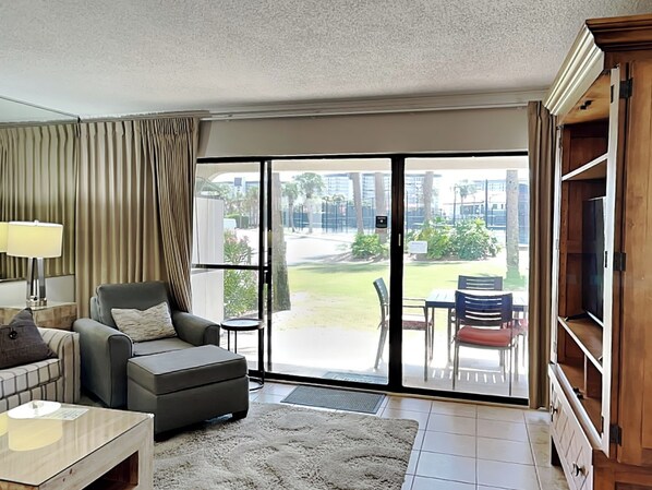 A living room with a sofa, armchair, and a coffee table. A sliding glass door leads to an outdoor area with a table and chairs.