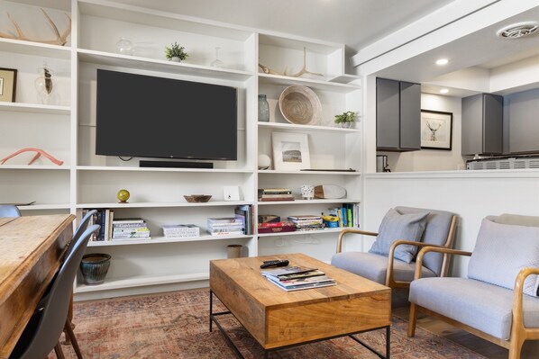 Un salon moderne doté d’un grand téléviseur à écran plat, d’une table basse en bois, d’un canapé gris et d’étagères blanches remplies de livres et d’objets décoratifs.