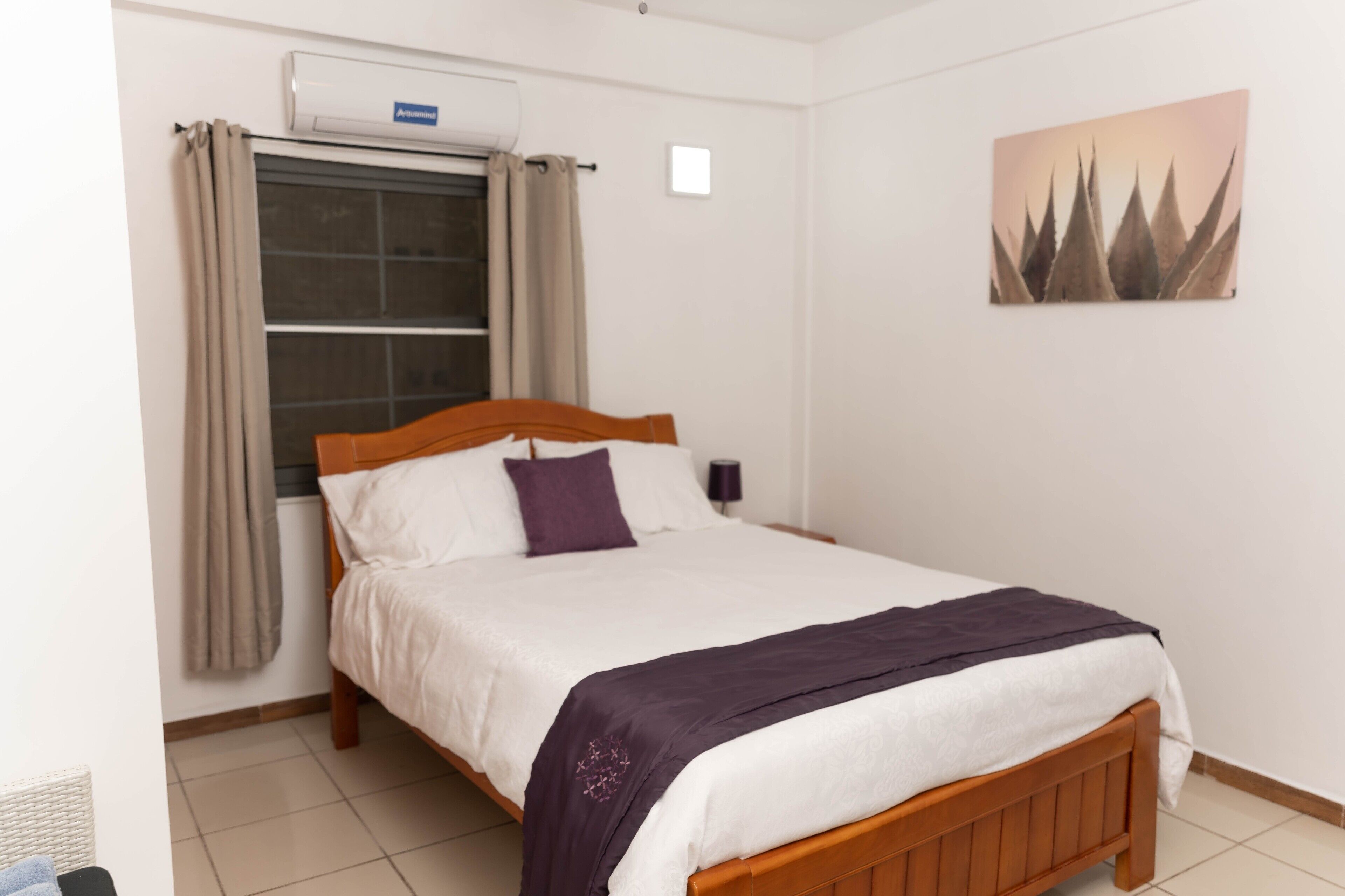 A neatly made bed with a wooden headboard, a window with curtains, and a wall-mounted air conditioner.