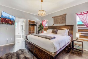 A bedroom with a large bed, a wooden headboard, a mounted TV, a bedside table with a lamp, and a view of the outside through a window. - 5BD – Large Yard & Firepit | Escape to the Mtns! (Cascade-Chipita Park)