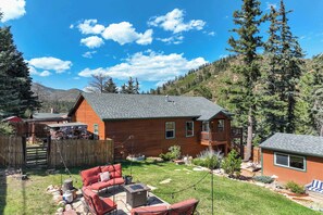 A wooden cabin with a fenced backyard, a patio with red furniture, and a mountain backdrop. - 5BD – Large Yard & Firepit | Escape to the Mtns! (Cascade-Chipita Park)