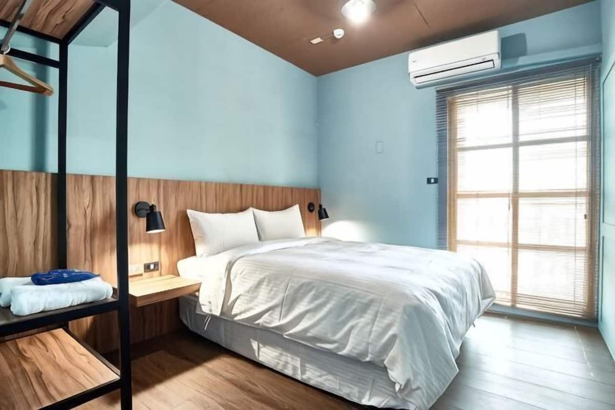 Ein Schlafzimmer mit Bett, Nachttisch, Lampe und einem Fenster mit Jalousien.