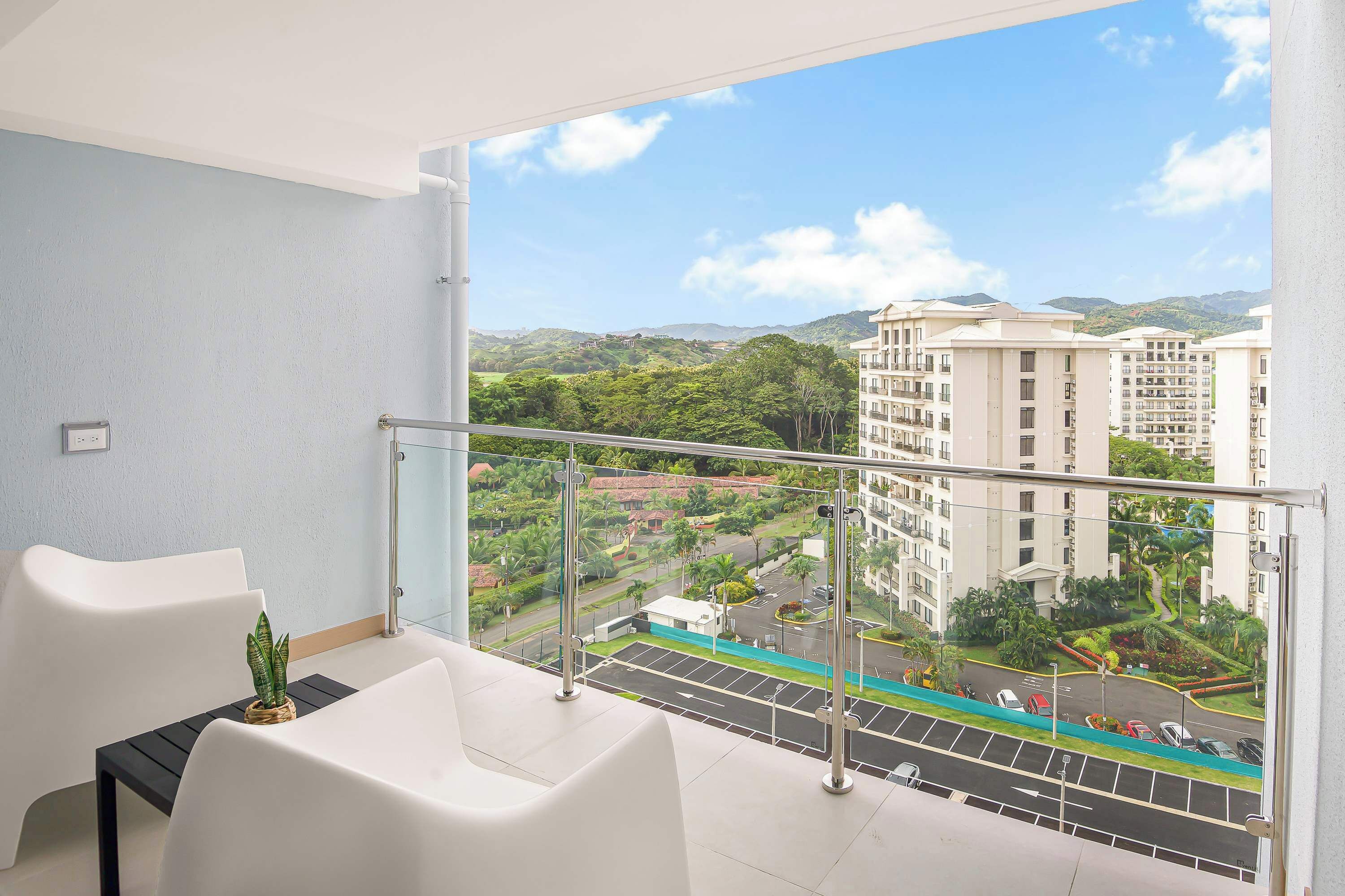 A balcony with white chairs, a small potted plant, and a view of a residential area with buildings and greenery.