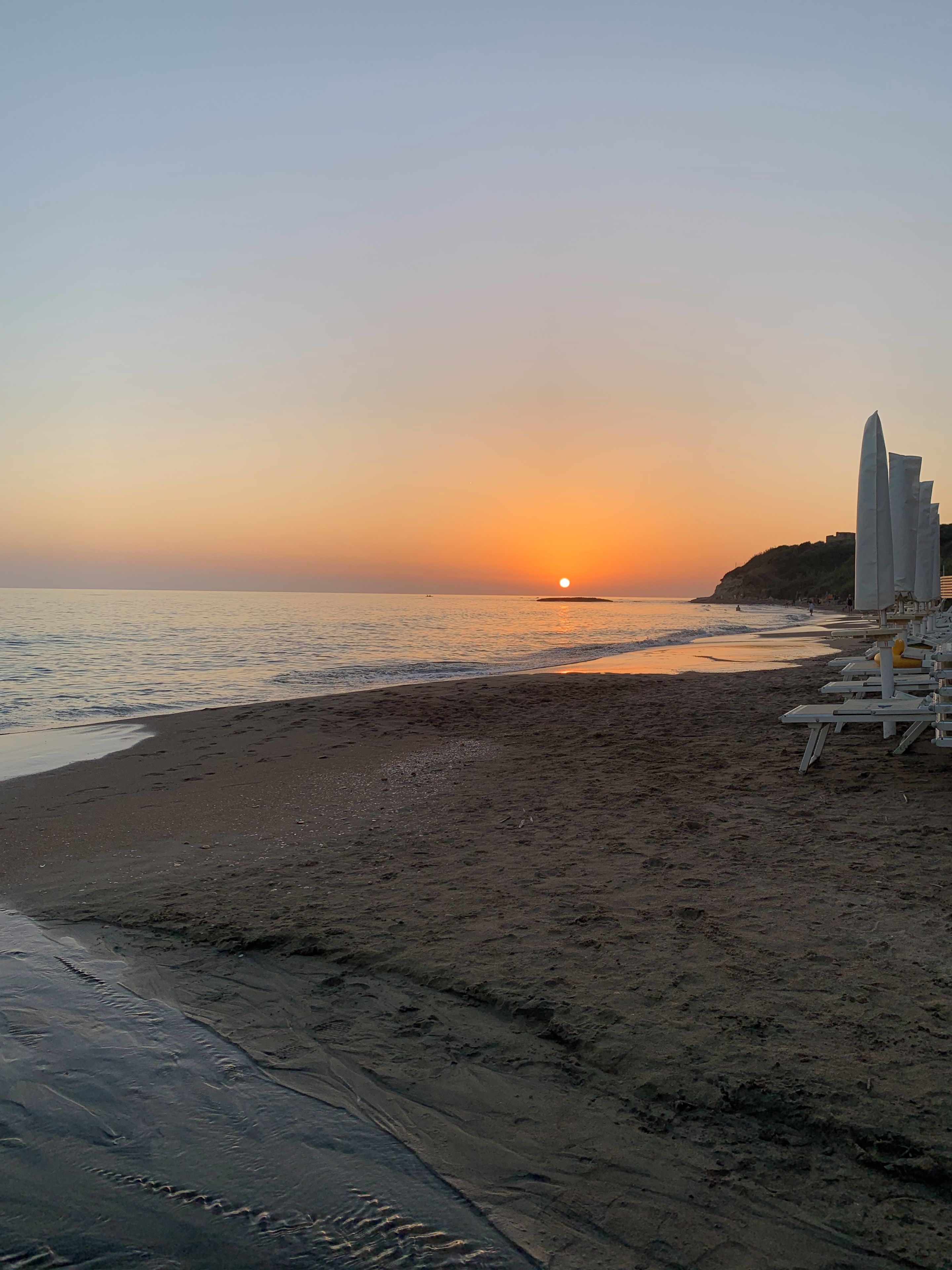 Une plage au coucher du soleil, un rivage sablonneux et une rangée de chaises longues blanches.