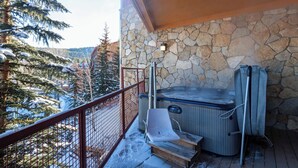 Un jacuzzi en un balcón con pared de piedra, baranda de madera y suelo cubierto de nieve.