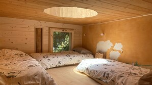Quarto acolhedor com paredes de madeira e uma grande janela, com duas camas e roupa de cama estampada, além de um mural de ovelhas na parede.
