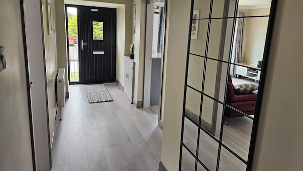 Un pasillo con una puerta negra, un tapete estampado y vistas a una habitación con una silla roja y un televisor.
