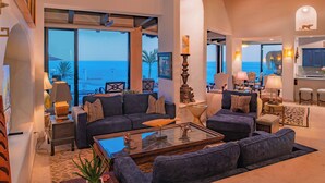 A spacious living room with a large window offering a view of the sea, a cozy seating area with sofas and a coffee table, and a kitchen area visible through an open doorway.