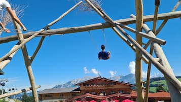 Altalena in legno con una persona che oscilla, un edificio sullo sfondo con ombrelloni rossi e montagne all'orizzonte.