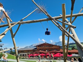 A wooden swing set with a person swinging, a building with red umbrellas in the background, and mountains in the distance.