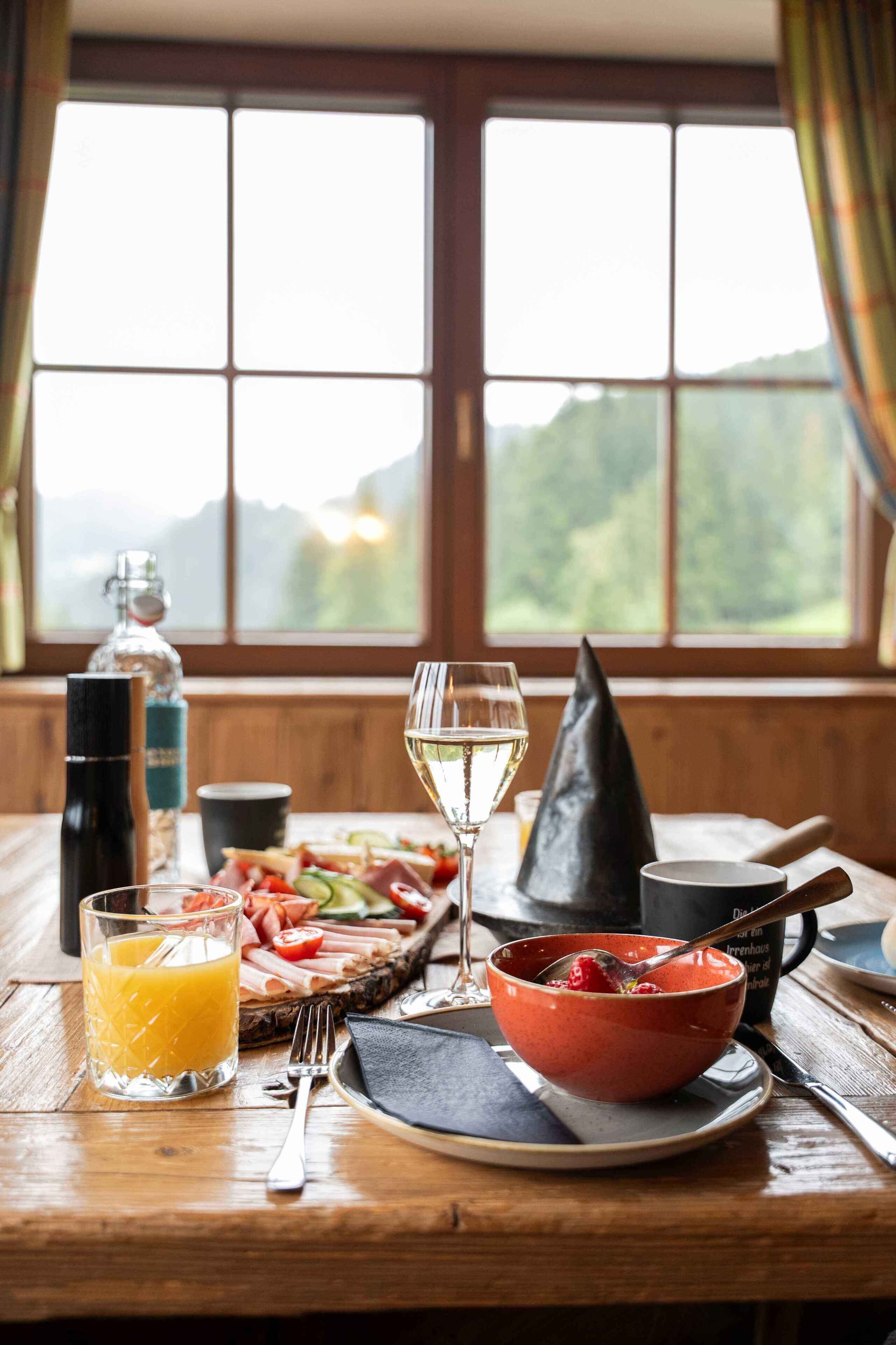 Ein Holztisch mit einer Mahlzeit, darunter ein Glas Weißwein, eine Schale rote Beeren, ein Teller mit Aufschnitt und ein Glas Orangensaft.