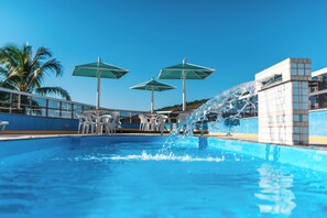 Una piscina con tres sombrillas, sillas blancas y una fuente de agua.