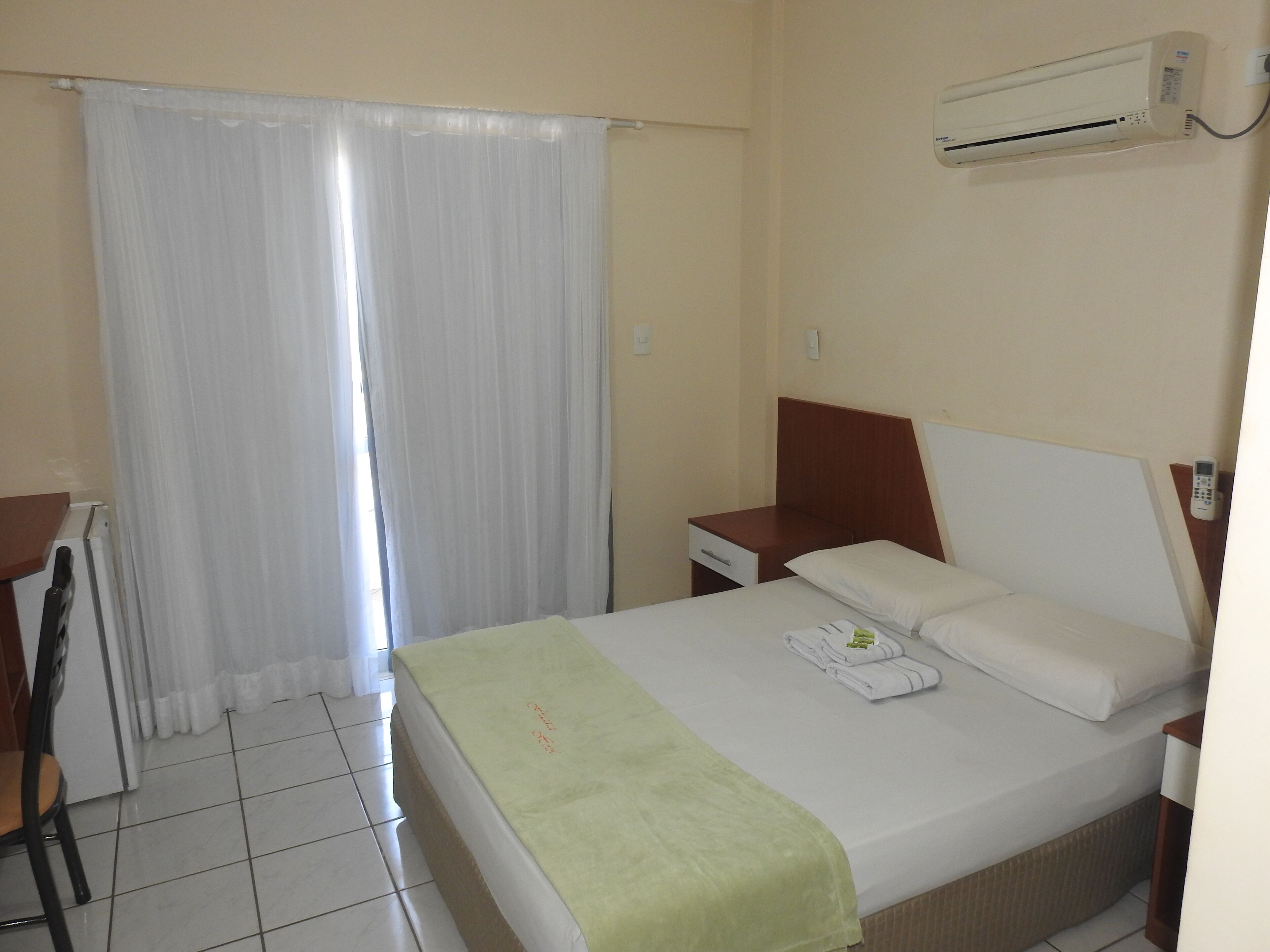 Habitación de hotel con cama, mesita de noche, una silla, ventana con cortinas e instalación de aire acondicionado.