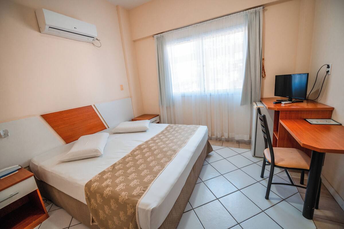 Una habitación de hotel con una cama, un escritorio con una computadora, una silla y un equipo de aire acondicionado.