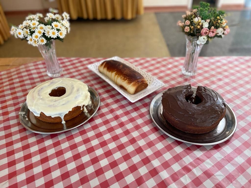 A table with a red and white checkered tablecloth, two cakes on metal plates, and two vases with flowers.