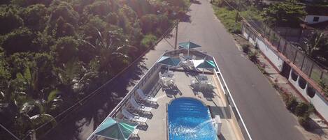 An aerial view of a rooftop pool area with lounge chairs and umbrellas, surrounded by greenery and overlooking a road.