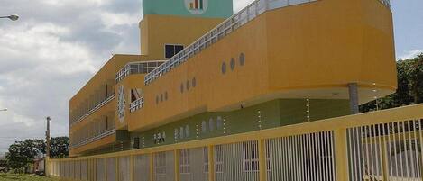 Un edificio moderno con fachada amarilla, una torre de reloj y un área cercada con plantas pequeñas.