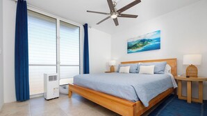 A bedroom with a bed, a ceiling fan, a heater, a window with curtains, a painting, and a nightstand with a lamp.