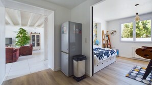 A modern, open-plan living space with a large window, a bed with a patterned bedspread, a dining area with a wooden table, and a built-in cabinet.