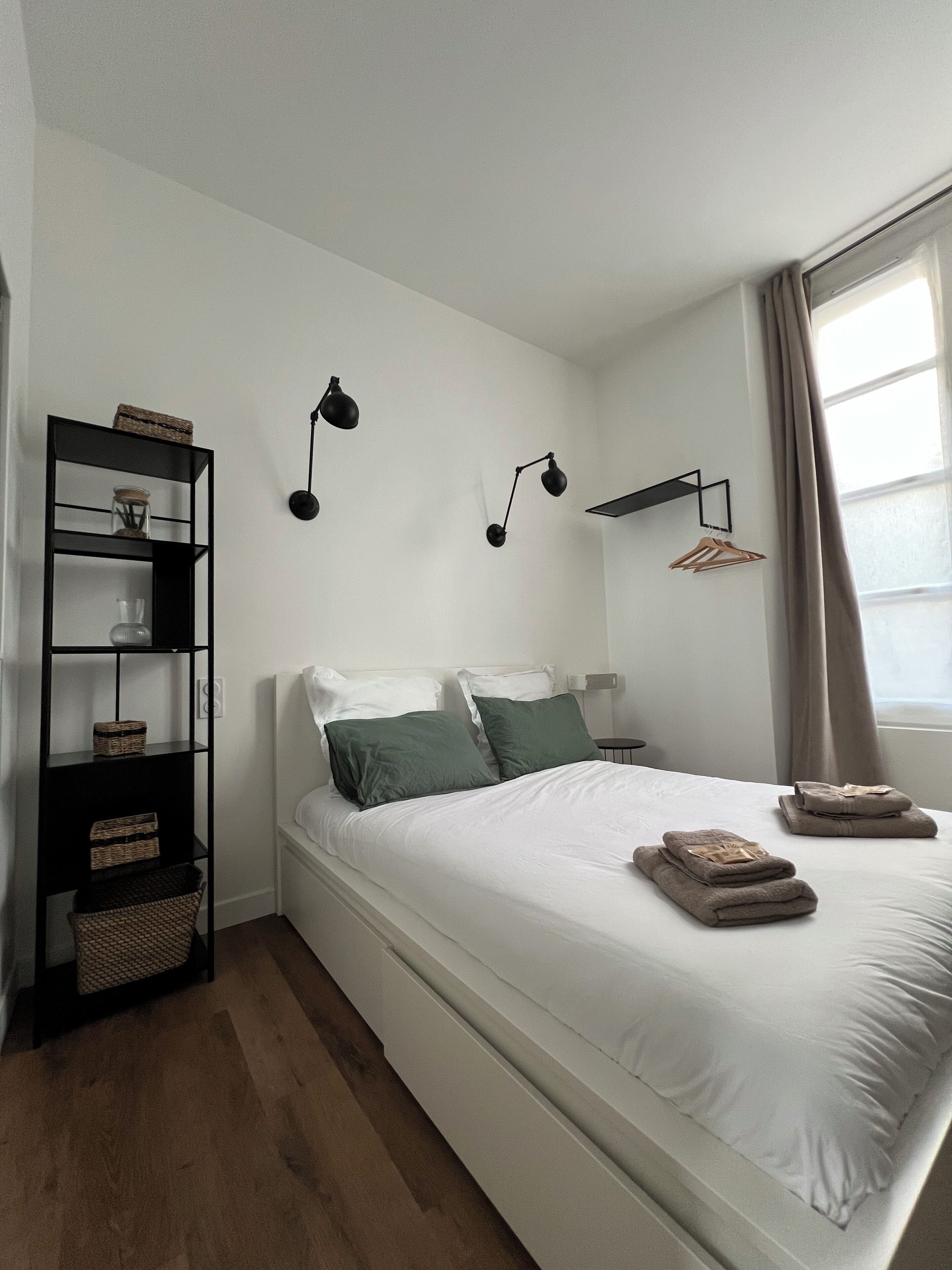 Een net opgemaakt bed met witte lakens en twee groene kussens, een zwarte boekenkast met manden, en twee wandlampen voor 's nachts te lezen.