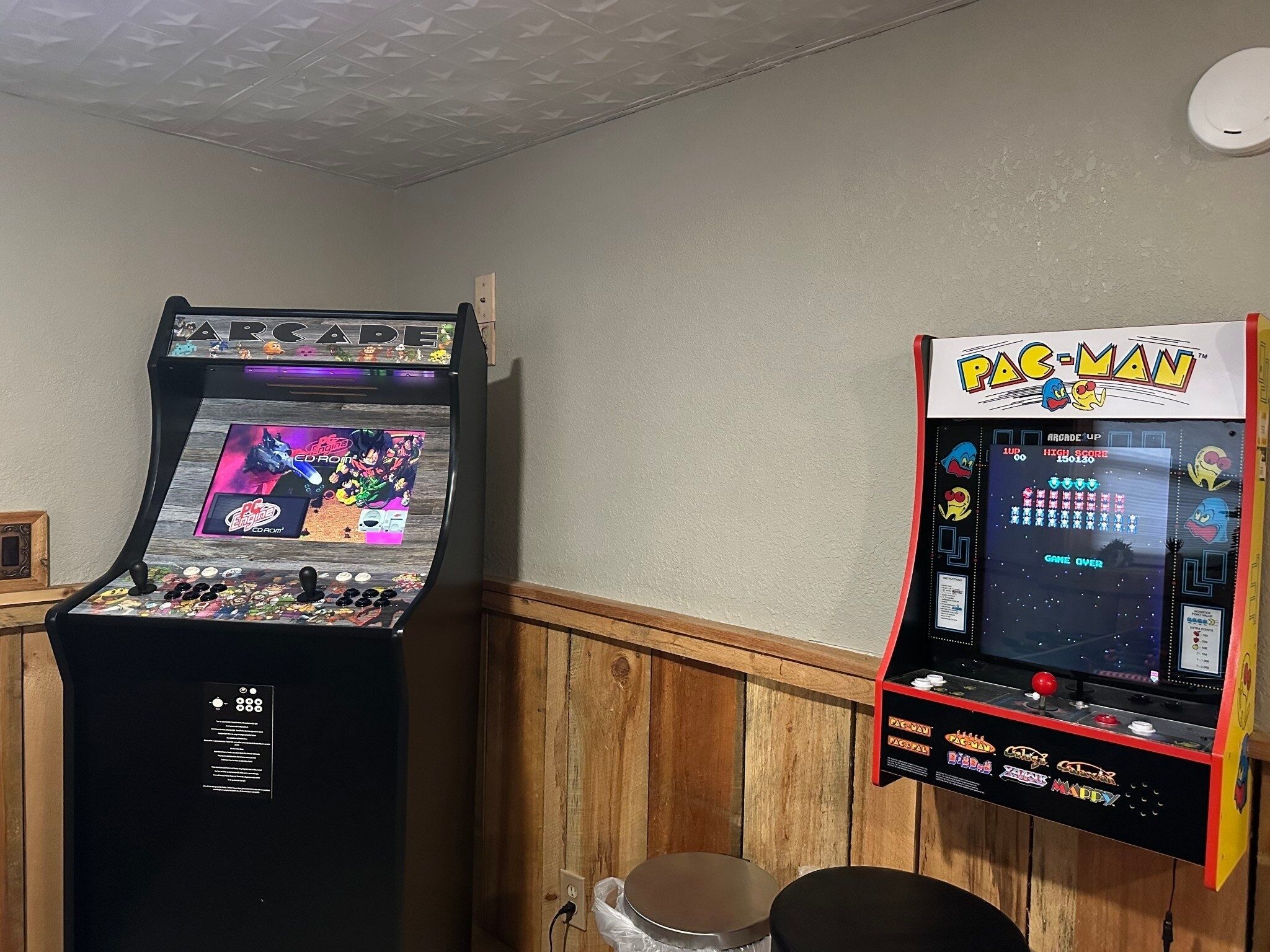 Two arcade machines, one displaying a fighting game and the other a classic arcade game, in a room with wooden paneling.