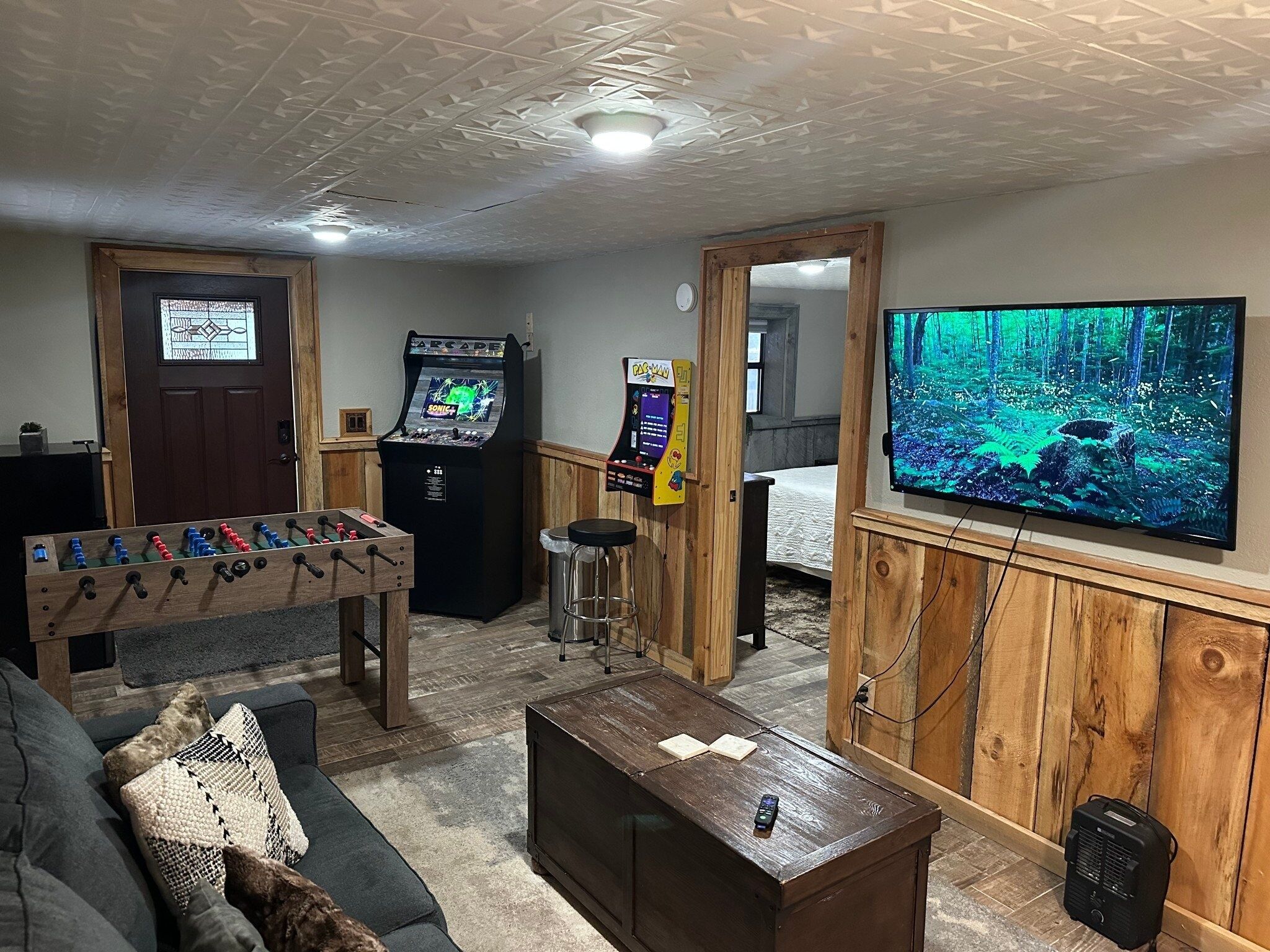 Una habitación con una mesa de fútbol de mesa, dos máquinas de videojuegos, un televisor que muestra un paisaje boscoso, un sofá y una mesa de centro de madera.