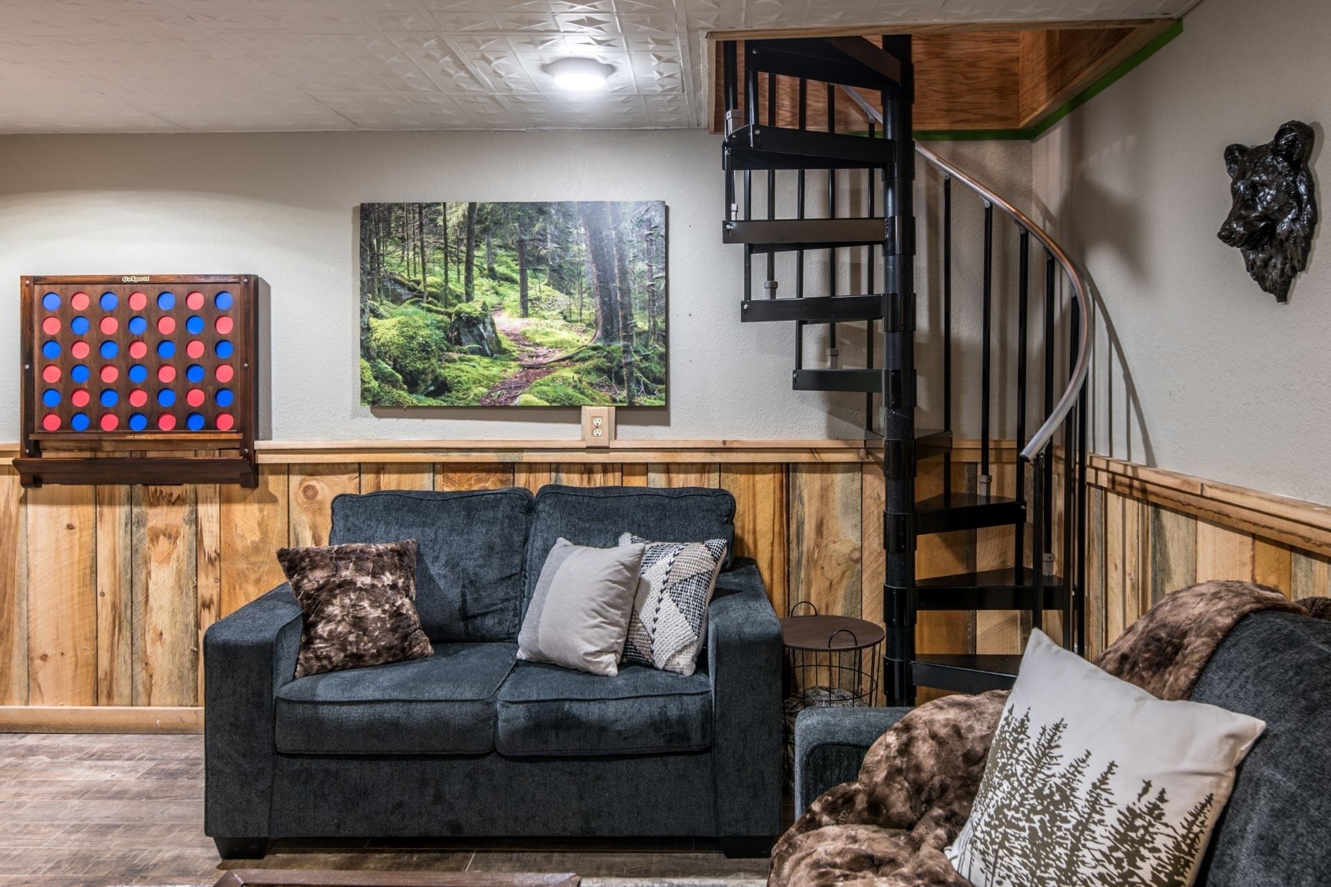 A living room with a dark blue sofa, a wooden accent wall, a painting of a forest, and a black spiral staircase.