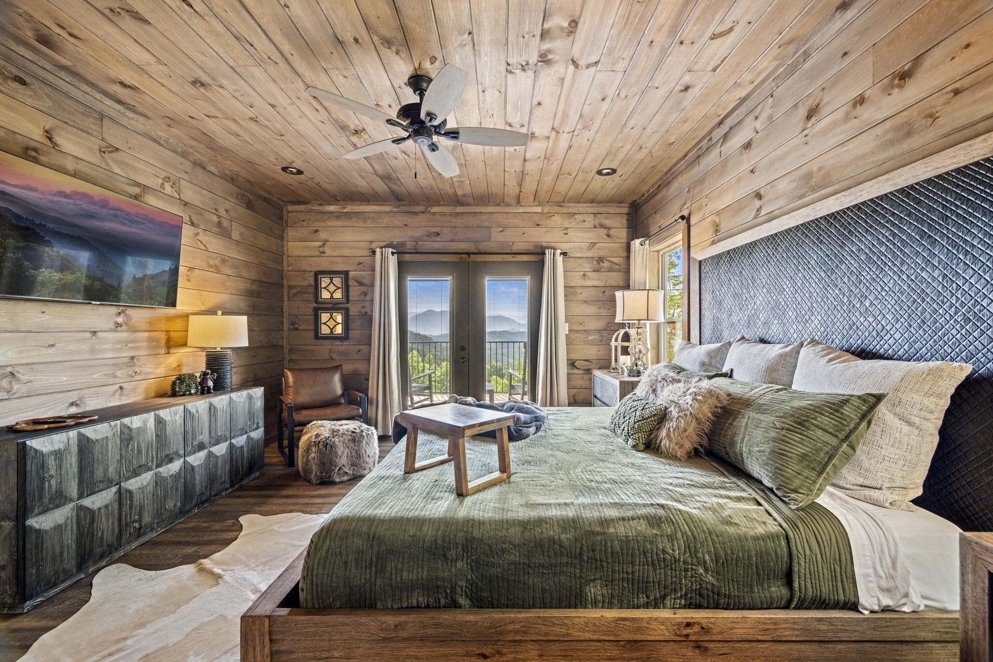 Acogedor dormitorio de cabaña de madera con cama, una mesita, ventilador de techo y vistas al exterior a través de una puerta.